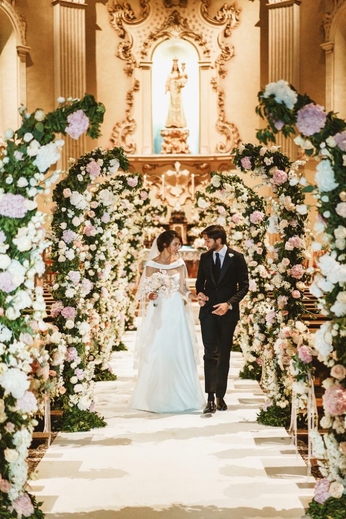 In questa foto una coppia di sposi mentre esce dalla chiesa per matrimonio. La navata centrale è decorata con semiarchi di ortensie bianche e rosa ed eucalipto ed edera rampicanti