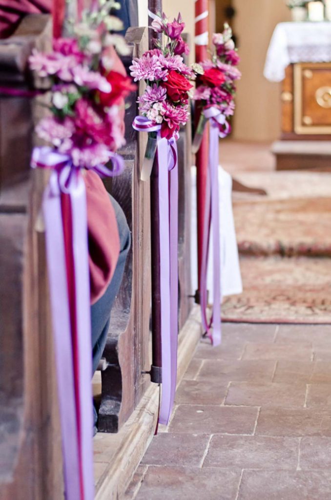 In questa foto 3 decorazioni floreali legate ai banchi di una chiesa. Le decorazioni sono realizzate margherite e fiori di campo colore lilla, rosa e magenta legate da 3 nastri abbinati e pendenti lungo il lato verticale dei banchi della chiesa. A destra si intravede l'altare e un tappeto