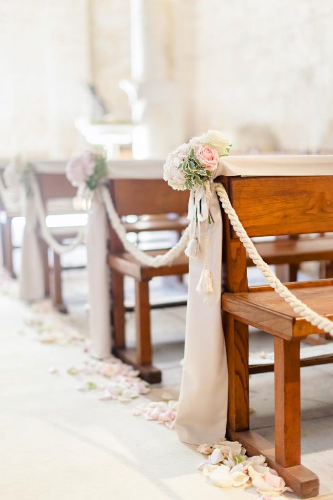 In questa foto addobbi chiesa per un matrimonio romantico ripresi da destra verso sinistra. I banchi sono decorati con drappi beige pendenti fino a a terra, mazzetti di fiori rosa e bianchi, legati da nappine beige. I banchi della chiesa sono infine collegate da un cordone beige. Lungo la navata sono sparsi petali di rosa bianchi