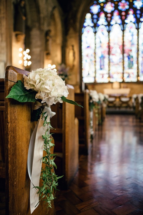 In questa foto il dettaglio di addobbi per matrimonio semplice. È mostrata una piccola composizione floreale legata ad un banco della chiesa in legno scuro, realizzata con un bocciolo di ortensia bianca, un ramoscello di foglioline e due nastri di raso grigio e rosa chiaro. Sullo sfondo si intravedono le altre composizioni legate ai banchi e l'altare con una grande vetrata posteriore di vetri colorati