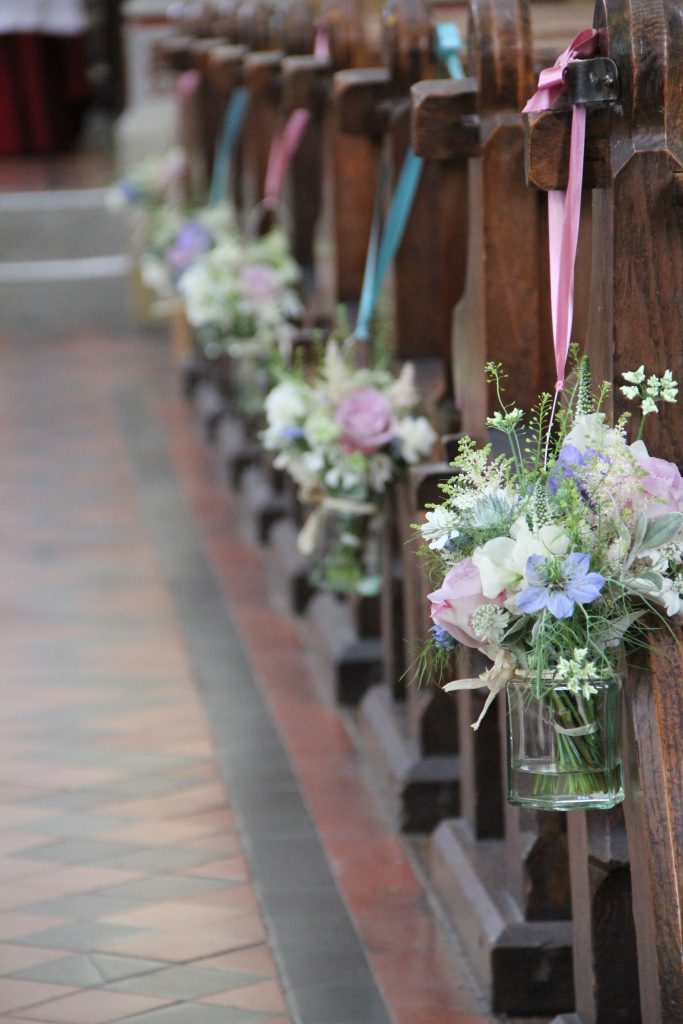 In questa foto il dettaglio di addobbi per chiesa matrimonio shabby chic. Ai banchi sono legati con nastri, alternati, azzurri e rosa, vasetti di vetro trasparente riempiti di acqua e fiori di campo lilla e bianchi, rose rosa e nebbiolina