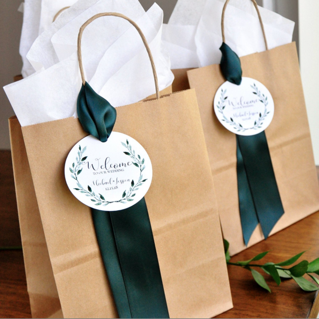 In questa foto due buste di carta marrone poggiate su un banco di una chiesa per matrimonio. Le wedding bag sono decorate con un nastro verde scuro e una targhetta rotonda con scritto "Welcome", i nomi degli sposi e la nata della nozze. Sul bordo della targhetta è dipinto un ramoscello di ulivo. Dalle buste fuoriescono fogli di carta bianca. 