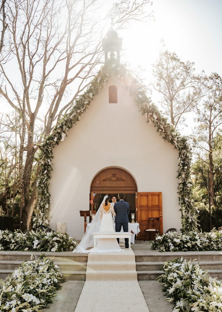 In questa foto 2 sposi in piedi davanti al sacerdote che celebra il loro matrimonio su un altare davanti al porto di ingresso di una chiesa di campagna. Il contorno della facciata della chiesa è interamente ricoperta d fiori bianchi e foglie rampicanti, usati anche nelle composizioni poste in cima ai 3 gradini che separa lo spazio per gli ospiti. Il corridoio centrale è ricreato con un tappeto bianco lungo i cui bordi, sul pavimento, sono presenti altre composizioni della stessa tipologia. Dietro la chiesa si vedono anche gli alberi alti di pino che la circondano da cui filtra la luce del sole 