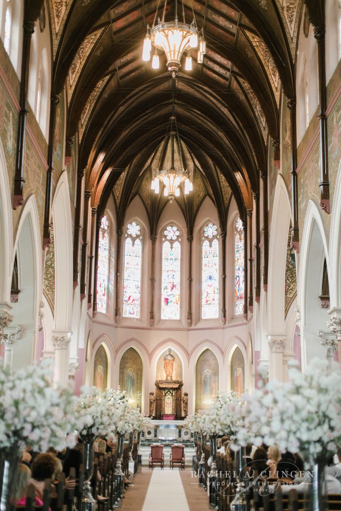 In questa foto una chiesa per matrimonio gotica con tetto in legno, colonne bianche e vetrate colorate. Lungo la navata sono disposti vasi alti e trasparenti con piccoli fiori bianchi e rosa. Alla fine della navata centrale si intravedono due poltrone rosa per gli sposi