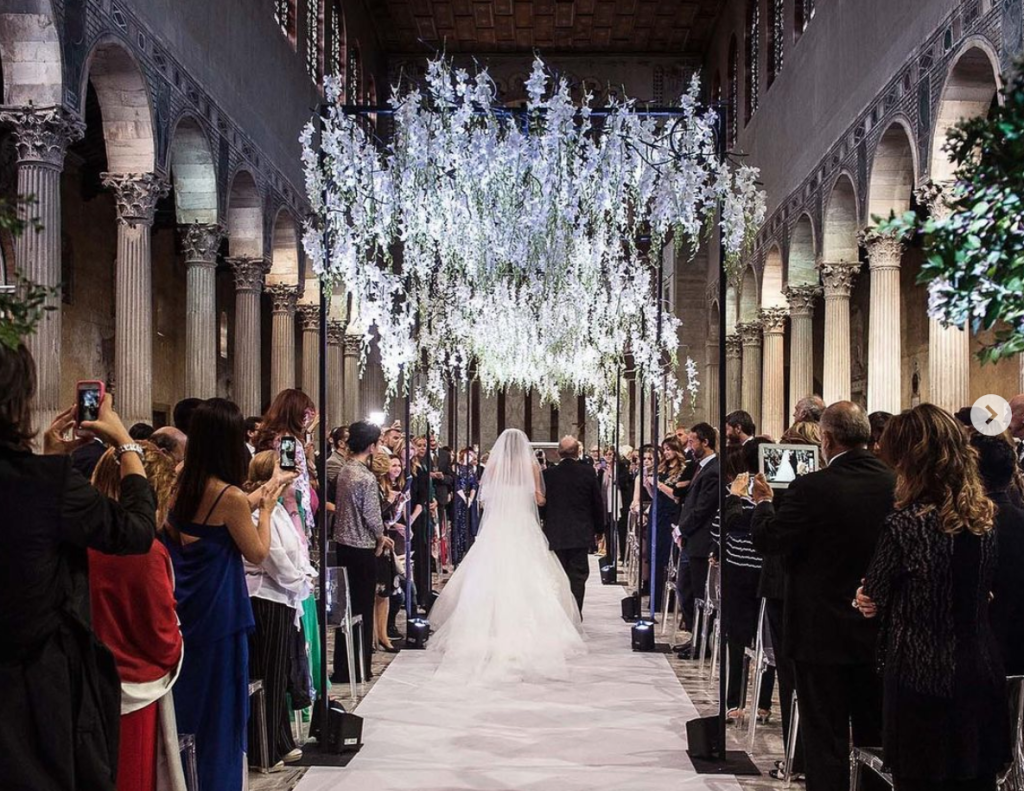 In questa foto l'interno di una chiesa durante un matrimonio. È ripreso l'ingresso della sposa che percorre la navata al braccio del padre mentre gli ospiti, in piedi e di spalle, li guardano e scattano foto con i loro smartphone. La navata è decorata con un tappeto bianco e a metà navata è presente un gazebo di gladioli bianchi e lilla illuminati. La chiesa è divisa in 3 navata da archi e colonne di marmo