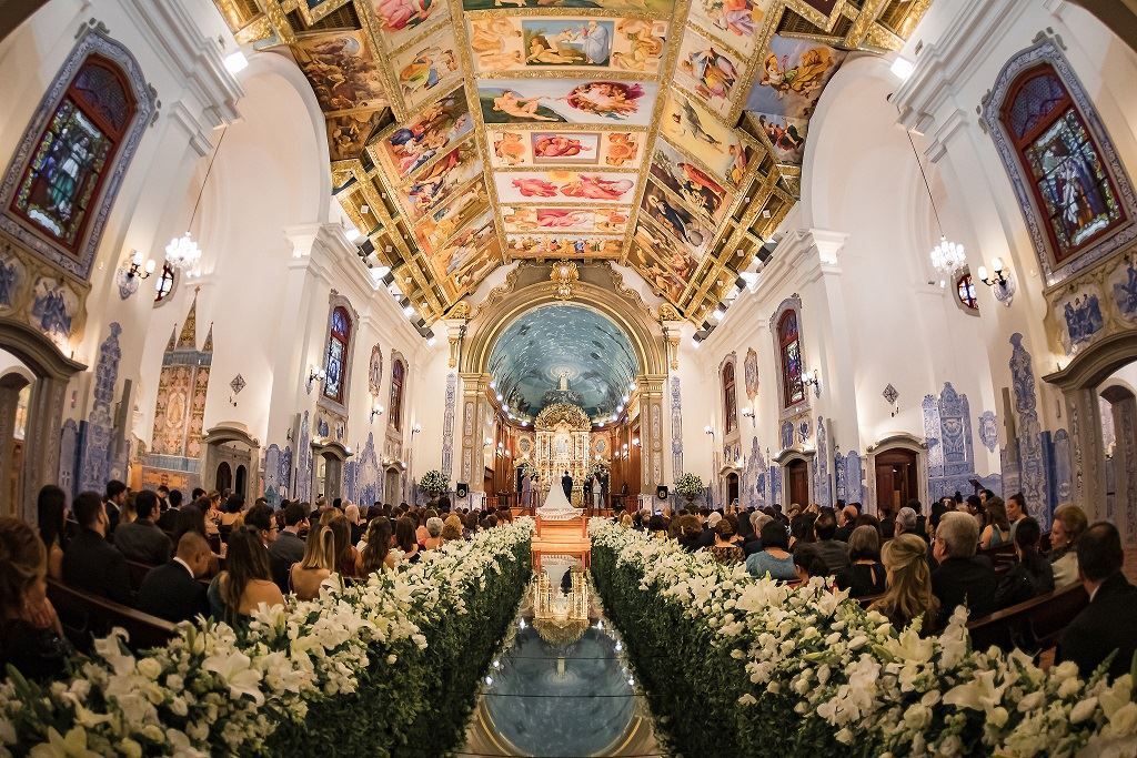 In questa foto una chiesa per matrimonio decorata con fiori bianchi lungo tutta la navata ricoperta da un tappeto riflettente. Ai lati sono seduti gli ospiti mentre sul fondo, all'altare, sono presente gli sposi in piedi. La chiesa presenta marmi grigi e azzurri lungo le pareti mentre il soffitto è ricoperto di dipinti e stucchi dorati. L'altare è coperto da una cupola azzurra