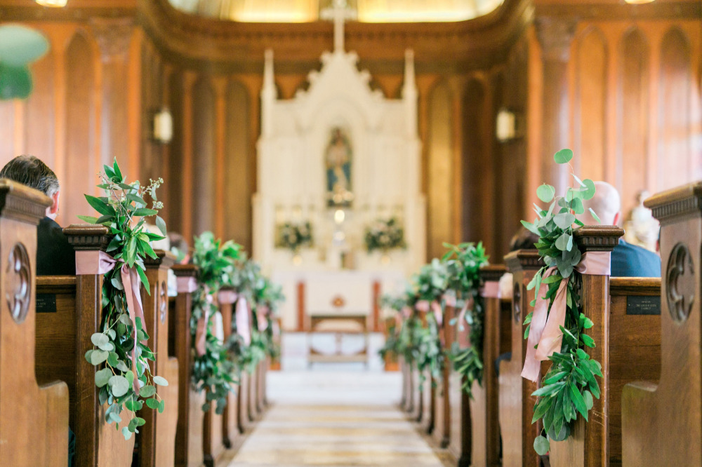 In questa foto una chiesa per matrimonio con pareti e banchi di legno decorati, a posti alternati, con mazzetti di eucalipto rivolti verso il basso fasciati da un nastro rosa cipria. Sullo sfondo si intravede l'altare bianco decorato con composizioni di foglie e fiori bianchi