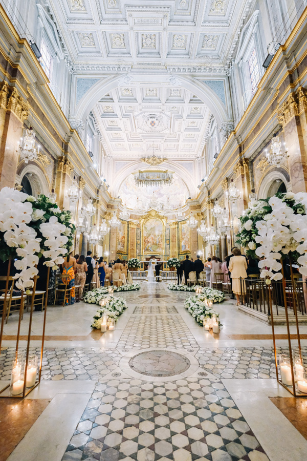 In questa foto una chiesa per matrimonio elegante ad archi di marmi e stucchi dorati e con tetto a volte e a cassettoni bianco e celeste. La chiesa è illuminata da lampadari in cristallo e candele. Sul fondo si intravedono gli sposi seduti, dietro all'altare 3 grandi dipinti religiosi. La navata è decorata con composizioni di orchidee poggiate su alzatine dorate a forma di parallelepipedo, con all'interno candele di diverse altezze. Composizioni di fiori bianchi e foglie sono disposte a forma circolare anche sul pavimento 