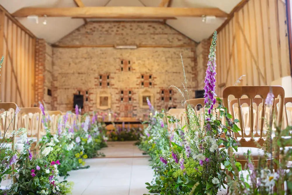 In questa foto una chiesa per matrimonio di campagna in legno decorata con composizioni di fiori di campo bianchi e viola e foglie verde poggiati su tutta la navata centrale fino a ricoprirla