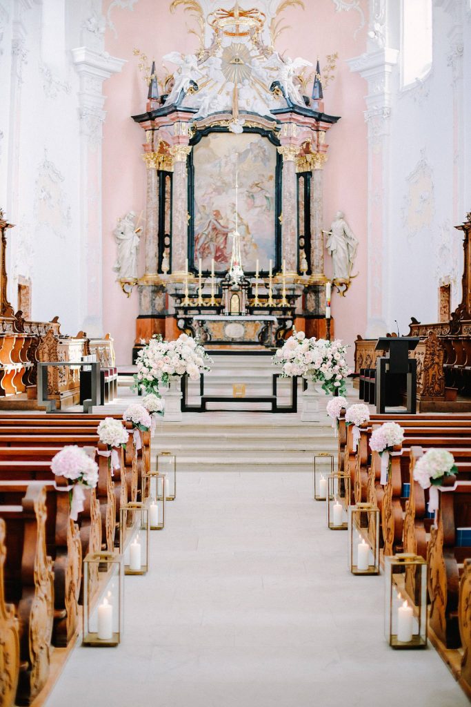 In questa foto una chiesa per matrimonio con pareti stuccate di bianco e altare rosa decorato con marmi neri e rosa e un grande dipinto dell'annunciazione. Ai banchi, a posti alternati, sono legati mazzetti di rose bianche, uguali a quelle usate per le decorazioni disposte sulla balaustra dell'altare. Lungo la navata, poggiate sul pavimento, sono disposti portacandele dorati a forma di parallelepipedo con all'interno candele grandi bianche accese