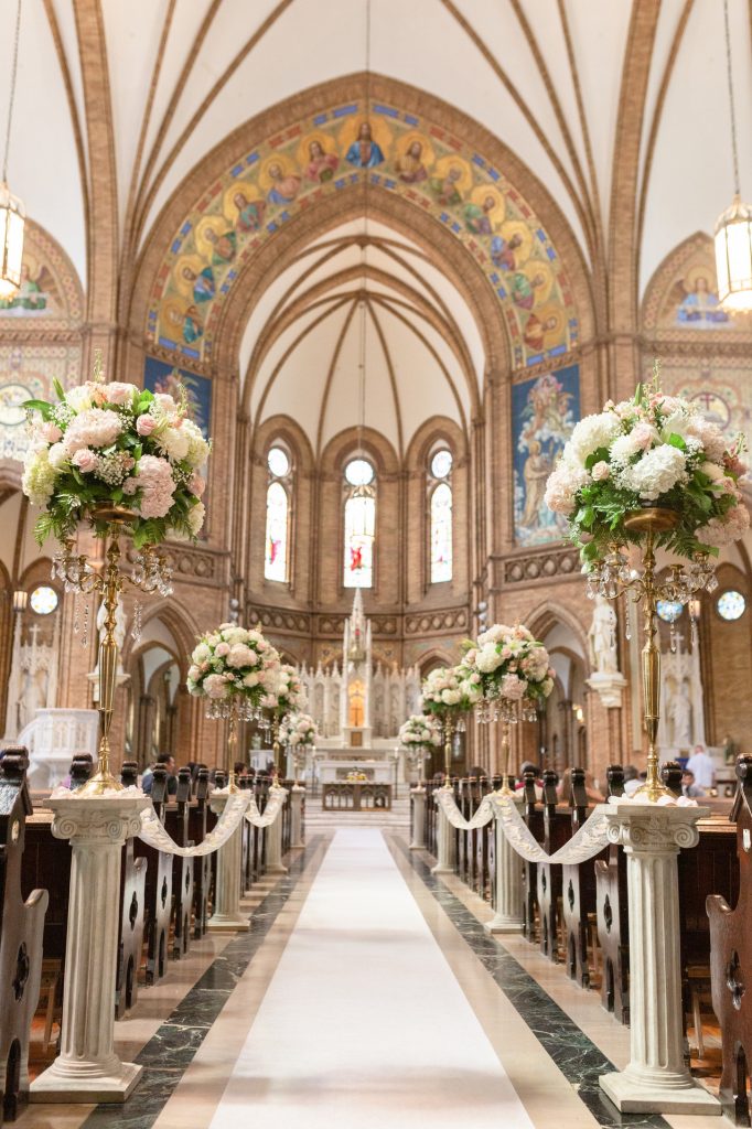 In questa foto una chiesa per matrimonio gotica con tetti a sesto acuto. La navata centrare è decorata con colonne greche di marmo su cui sono poggiate alzatine dorata con composizioni rotonde di fiori rosa e bianchi e foglie. Le colonne sono collegate da larghi nastri di pizzo ricamato. Sopra l'altare sono presenti dei mosaici bizantini dorati che raffigurano santi