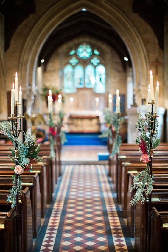 In questa foto una chiesa per matrimonio gotica con archi a sesto acuto in marmo grigio e pavimento a quadri bianchi, bordeaux e blu. La navata è decorata con candelabri dorati lunghi con 3 candele accese e poste a diverse altezze. Sulle aste dei candelabri sono attorncigliati dei ramoscelli di pino, rose rosa e fiori rossi