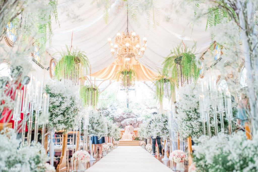 In questa una chiesa per matrimonio decorata con tende bianche sul soffitto e immersa in addobbi floreali di fiori bianchi lungo tutta la navata. Dal soffitto pendono composizioni di gelci. Sullo sfondo si intravedono gli sposi davanti ad un altare di pietra e un grande crocifisso che lo sovrasta