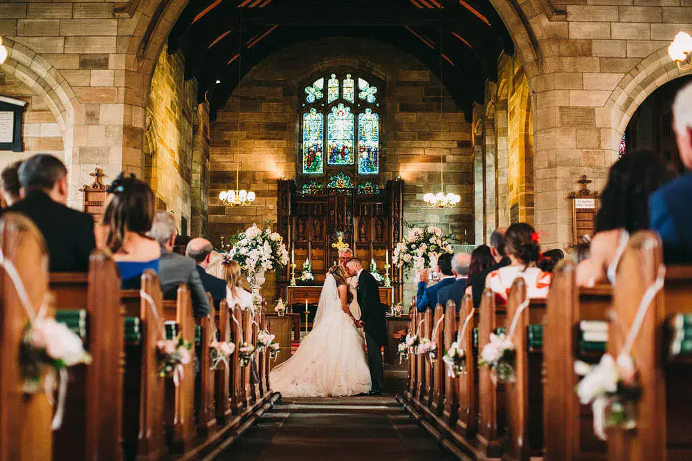 In questa foto due sposi si baciano alla fine del loro matrimonio in chiesa davanti al sacerdote ripresi da metà navata. I loro ospiti seduti di spalle li guardano. Legati ai banchi della chiesa, a posti alternati lungo la navata, sono appesi devi piccoli vasetti con dentro acqua e mazzetti di fiori bianchi e rosa. Le composizioni sono completate da nastri bianchi e rosa