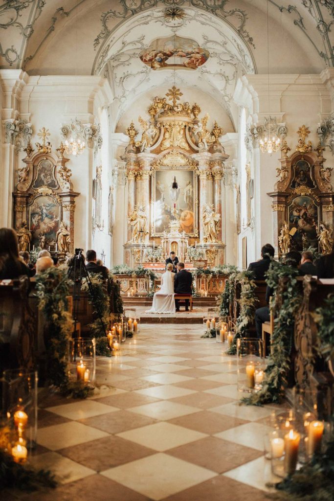 In questa foto un matrimonio in una chiesa barocca con pareti bianche, stucchi dorati e marmi all'altare e sulle nicchie laterali. All'altare sono presenti gli sposi seduti e dietro, ai banchi, i loro ospiti. La navata con grandi mattoni a forma di rombo di marmo rosa e bianco è decorata con composizioni di foglie rampicanti sui banchi mentre alla base sono disposti portacandele cilindrici con candele di diverse altezze accese