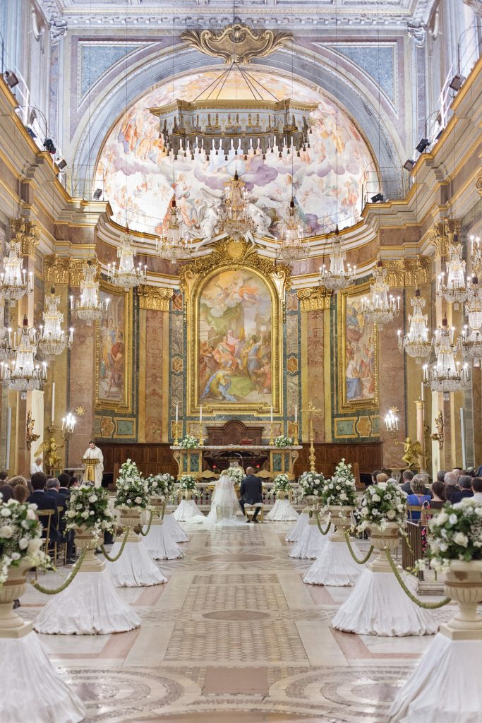 In questa foto un matrimonio in una grande chiesa decorata con addobbi eleganti. Lungo la navata sono disposte alzatine ricoperte di stoffa bianca su cui sono poggiati vadi di marmo con composizioni di fiori bianchi e foglie. I vasi sono legati l'uno all'altro da cordoni spessi di colore verde. Alla fine della navata ci sono 2 sposi seduti davanti al sacerdote. Ai lati, oltre il cordone di fiori, sono disposti gli ospiti. La chiesa è riccamente decorata con diversi lampadari di cristallo mentre sull'altare sono presenti grandi dipinti sacri che ricoprono interamente la parete