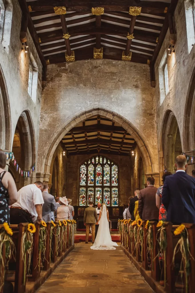 In questa foto una chiesa durante un matrimonio con archi in marmo e tetto in legno. La navata centrale è decorata con girasoli e nastri verdi e bianchi legati ai bianchi. Ai lati della navata, tra i banchi ma in piedi sono presenti gli ospiti delle nozze. In fondo alla navata, in piedi, gli sposi si tengono per mano davanti al sacerdote. In fondo all'altare è presente una vetrata colorata