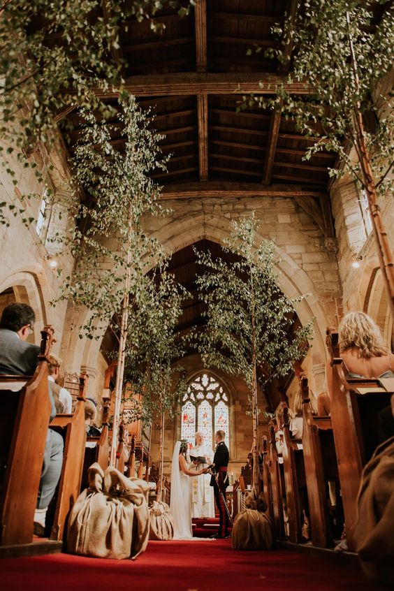 In questa foto un matrimonio in una chiesa gotica in pietra con tetto in legno ripresa dal basso verso l'alto. All'altare due sposi si tengono per mano mentre recitano le promesse davanti al sacerdote mentre i loro invitati ascoltano seduti. La navata è decorata con un tappeto rosso alberi alti d'ulivo con i vasi chiusi in sacchetti di iuta 