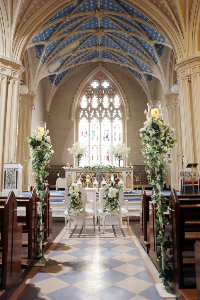 In questa foto una chiesa per matrimonio con pareti in marmo bianco e tetto azzurro con stelle. È ripreso l'altare da metà navata decorata con portacandele con rami di edera rampicanti e fiori bianchi. Sulla mensa dell'altare sono presenti due alberelli di fiori bianchi e una decorazione rampicante. Al centro del navata sono presenti le sedie bianche per gli sposi con composizioni di fiori bianchi e foglie sullo schienale. Davanti sono presenti gli inginocchiatoi bianchi