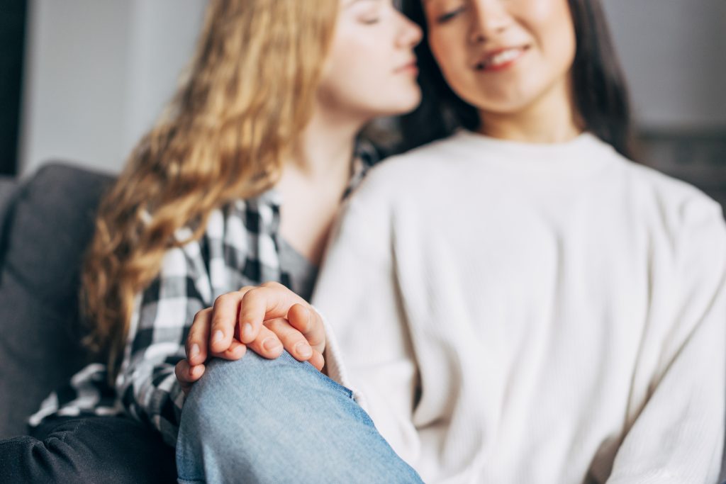 In questa foto due ragazze sul divano si stringono le mani poggiate sulle gambe. La ragazza a sinistra si avvicina all'altra come per baciarla sulla guancia