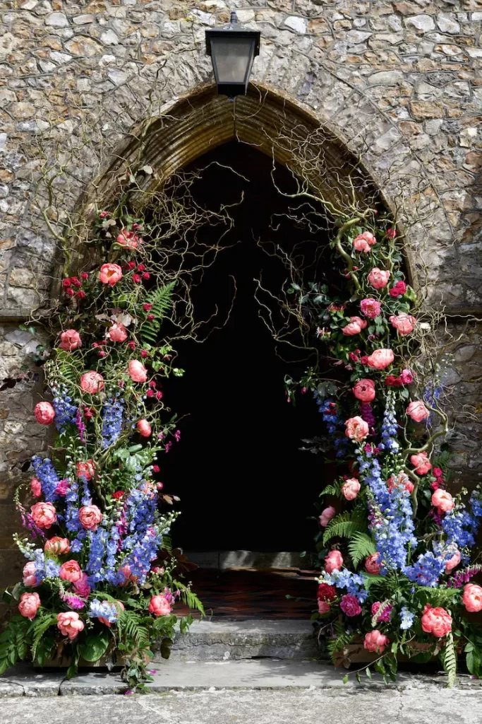 In questa foto l'arco in pietra di chiesa di campagna addobbato per un matrimonio. Su tutto l'arco sono disposti fiori rampicanti lilla, rosa e fuxia e foglie