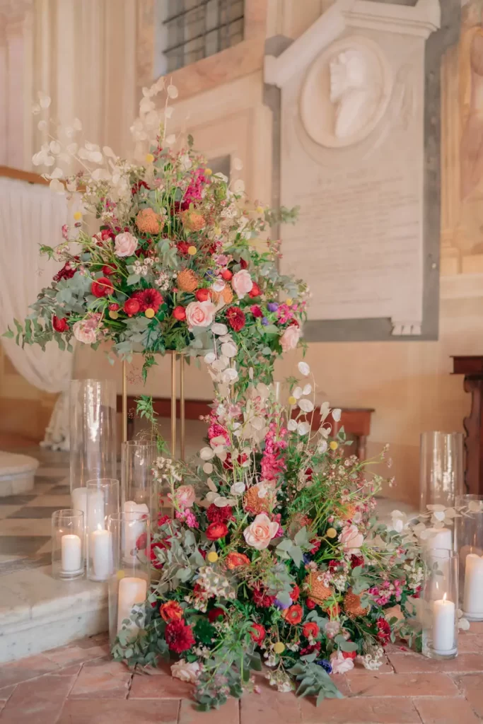 In questa foto due composizioni di fiori all'angolo dell'altare di una chiesa realizzate con fiori rossi, arancioni, rosa e fuxia. Una composizione è poggiata sul pavimento sotto un gradino di marmo. L'altra composizione è poggiata su un'alzatina dorata a forma di parallelepipedo sopra il gradino. Entrambe sono circondate da candale bianche di varie altezze contenute in portacandele di vetro trasparente a forma cilindrica