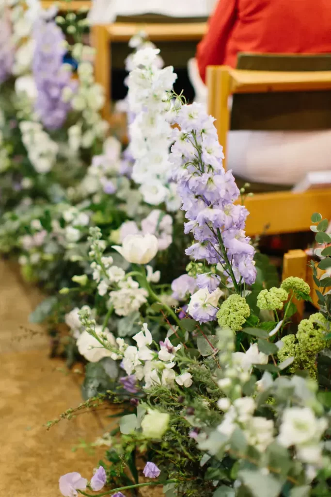 In questa foto una fila di composizioni  floreali poggiate lungo la navata accanto ai banchi di una chiesa. I fiori usati solo gladioli colore lilla e bianco e rose bianche intrecciati a foglie e arbusti