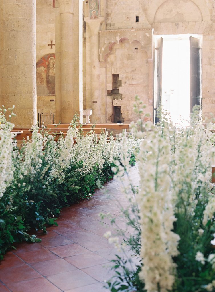 In questa foto una chiesa rustica con pareti e colonne in pietra, decorata per un matrimonio. La navata centrale è interamente ricoperta sui lati di composizioni di foglie e gladioli bianchi