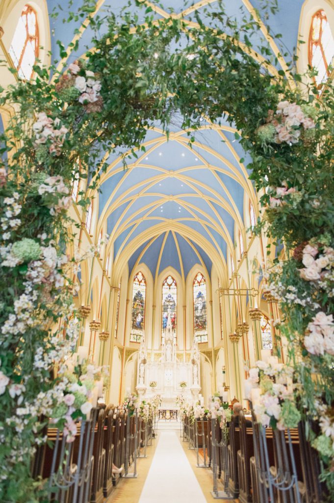 In questa foto una chiesa decorata per un matrimonio religioso. In primo piano è presente un grande arco di foglie e fiori rosa e bianchi. Gli stessi fiori decorano i banchi della chiesa. Dall'arco di fiori si intravede il tetto ad archi azzurro e l'altare con vetrate a mosaico in stile gotico