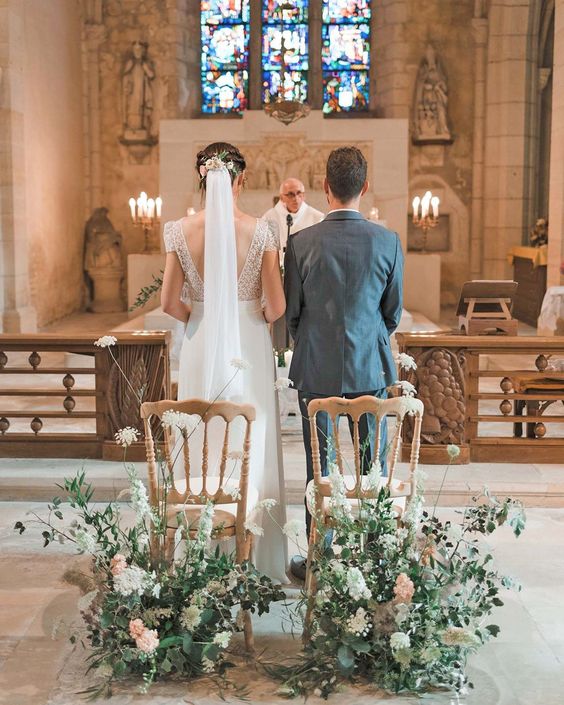 In questa foto due sposi di spalle in piedi durante la celebrazione del loro matrimonio religioso in chiesa mentre il sacerdote recita l'omelia. Le indossa un velo di media lunghezza fermato sulla testa da una piccola corona di fiori rosa e bianchi, gli stessi fiori usati come decorazioni alla base delle sedie chiavarine colore oro.