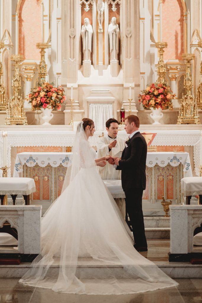 In questa foto due sposi si guardano e si tengono le mani mentre si scambiano le fedi in chiesa durante il loro matrimonio religioso. Il sacerdote regge il microfono allo sposo ripetendo le promesse. In primo piano è mostrato il velo disteso tra lo spazio dell'altare e il marmo della navata centrale. Dietro di loro un altare di marmo rosa e bianco