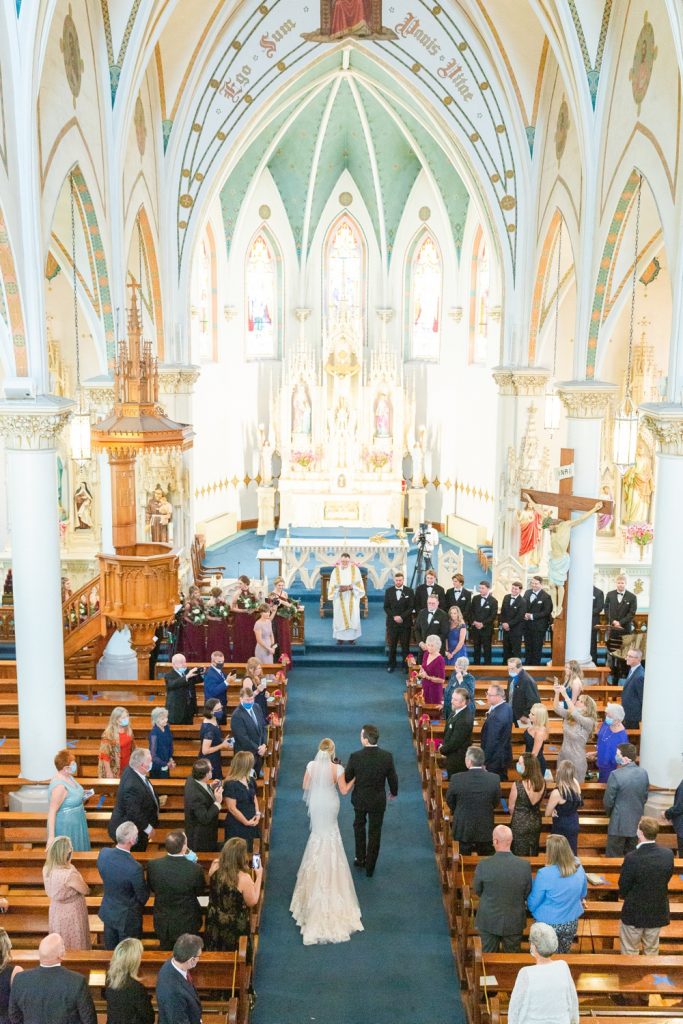 In questa foto l'ingresso della sposa in chiesa al braccio del padre, ripreso dall'alto. Sposa, papà ed ospiti ai banchi sono ripresi di spalle. Ad aspettarli all'altare il sacerdote che celebrerà la messa, i testimoni e le damigelle in abito bordeaux. La navata è ricoperta da un tappeto blu navy