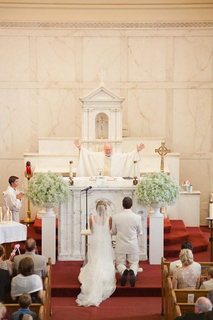 In questa foto due sposi inginocchiati all'altare davanti al sacerdote che li benedice a braccia aperte