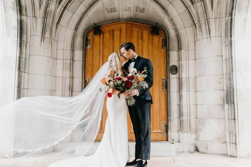 In questa foto due sposi si baciano davanti alla porta di una chiesa gotica in marmo e legno. Il velo della sposa svolazza. Entrambi gli sposi tengono tra le mani il bouquet di fiori rossi, rosa, gialli e foglie verdi