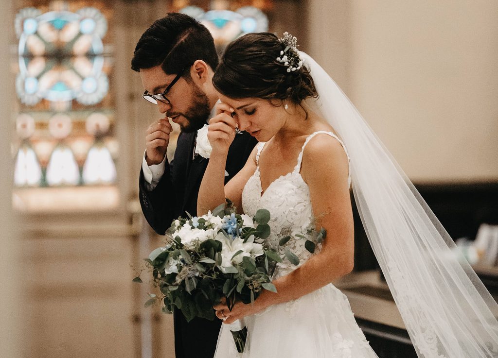 In questa foto due sposi si fanno il segno della Croce durante una preghiera del loro matrimonio in chiesa. Entrambi gli sposi hanno gli occhi chiusi e la testa abbassata. In primo piano è mostrato il bouquet di alloro, fiori bianchi e blu.