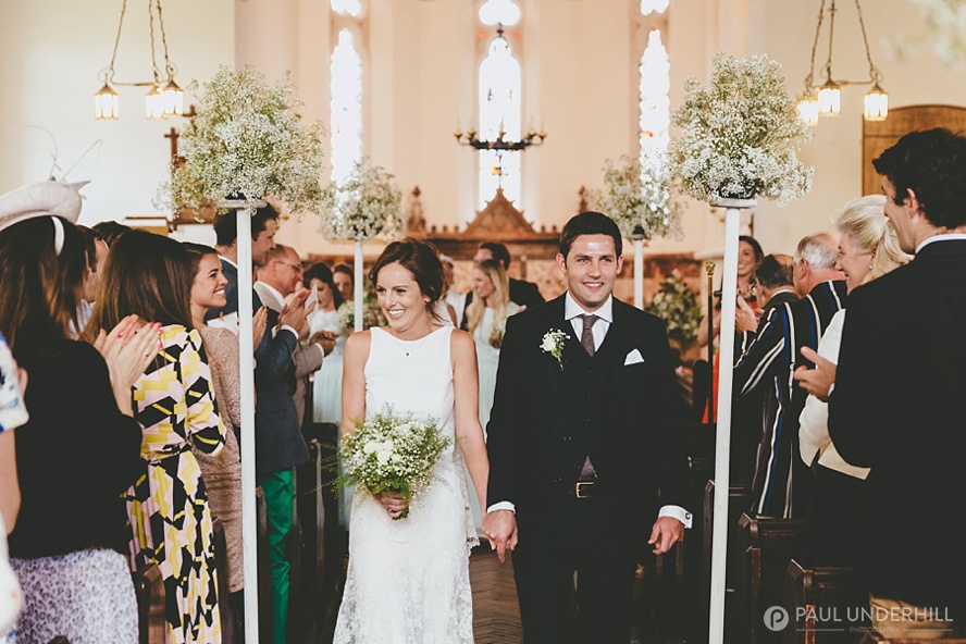 in questa foto due sposi mentre escono dalla chiesa dopo il loro matrimonio. Li applaudono gli ospiti ripresi di spalle, la navata è decorata con sottili colonne bianche sulla cui cima sono presenti composizioni di fiori velo di sposa, abbinati al piccolo bouquet che la sposa tiene nella mano destra e alla boutonnière dello sposo. Dietro si intravedono le damigelle vestite con abiti verde menta