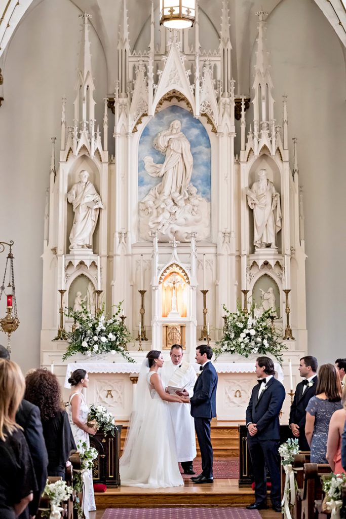 In questo articolo due sposi si tengono per mano all'altare durante la celebrazione del loro matrimonio religioso. Il sacerdote recita la benedizione mentre tutti gli ospiti assistono alla cerimonia. La chiesa è decorata con statue di marmo bianco all'altare e con fiori bianchi lungo la navata e all'altare