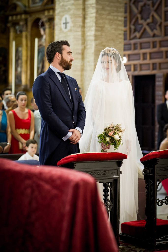 In questa foto due sposi ripresi in obliquo si guardano innamorati all'altare prima del loro matrimonio religioso. Lei è ancora interamente coperta dal suo velo cattedrale  ma si intravede un diadema tra i capelli e tiene nella mano destra un piccolo bouquet di fiori di campo. Lo sposo indossa un tight blu navy. Davanti a loro degli inginocchiatoi di legno scuro e velluto rosso porpora. Dietro di loro si intravedono alcuni ospiti