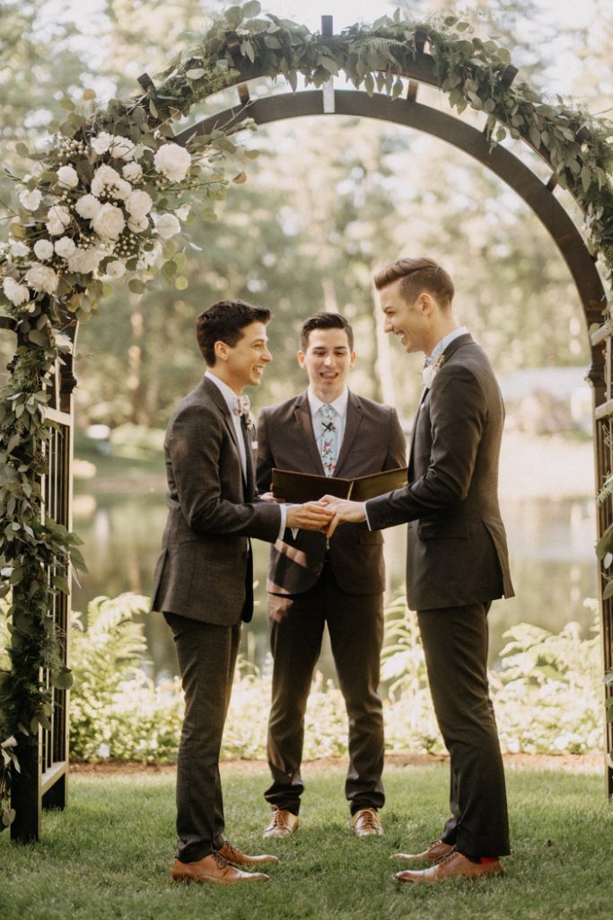 In questa foto due sposi ridono tenendosi per mano durante il loro matrimonio gay in un parco e sotto un arco di foglie e rose bianche. Dietro di loro ad officiare un giovane ufficiale di stato civile