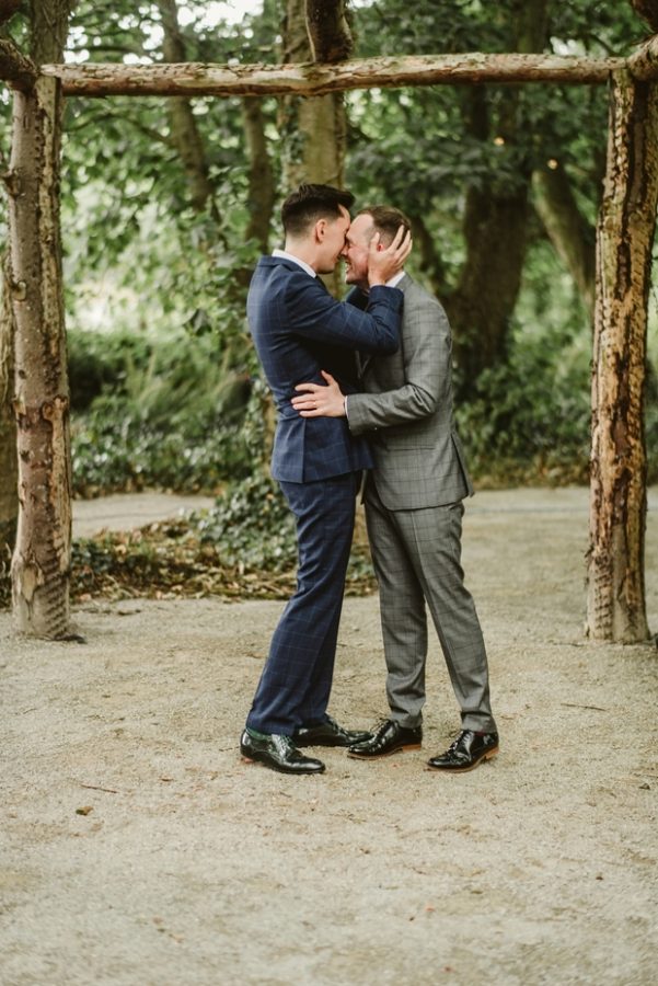 In questa foto due sposi gay alla fine della cerimonia della loro unione civile in un bosco si abbracciano davanti ad un arco di legno. Lo sposo a sinistra indossa un abito blu a quadri e stringe tra le mani la testa dell'altro sposo a destra in abito grigio a quadri che gli cinge la vita con le mani