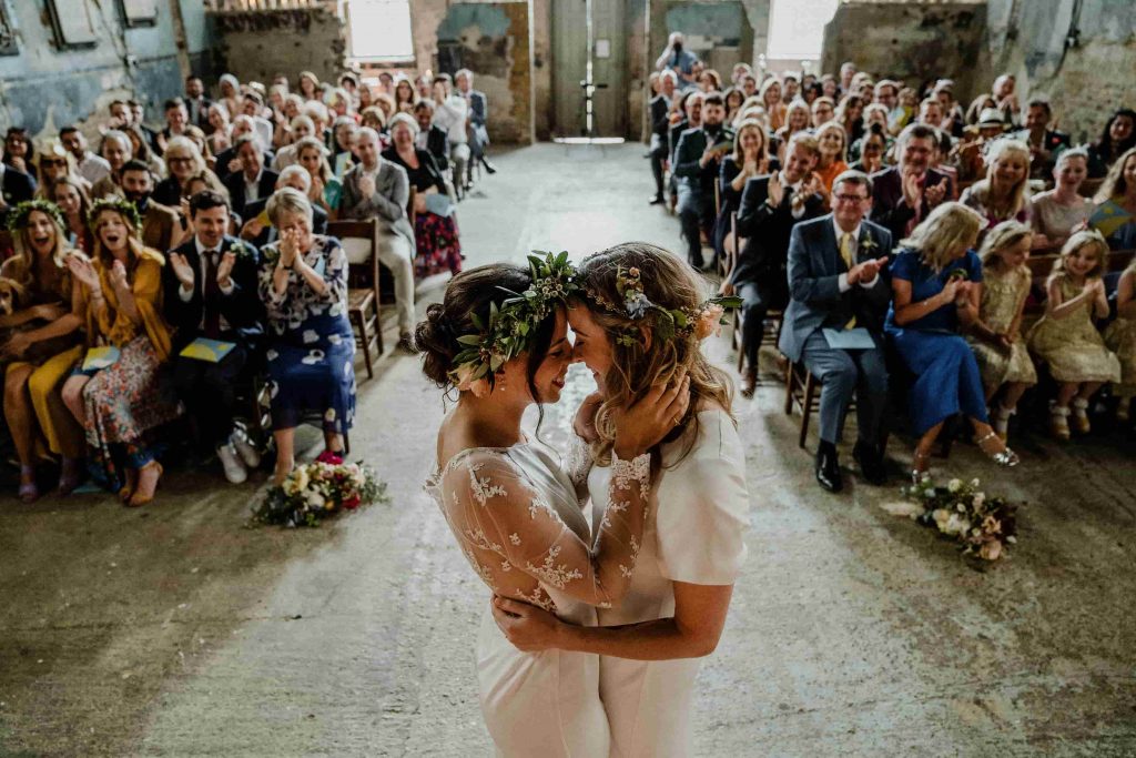 In questa foto due spose felici si guardano durante la cerimonia della loro unione civile in una chiesa sconsacrata mentre i loro ospiti applaudono e festeggiano. Entrambe le spose indossa delle coroncine di fiori e foglie. La sposa a destra è mora con i capelli raccolti e tiene le pani sul collo della sua compagna bionda con i capelli sciolti che la cinge sui fianchi