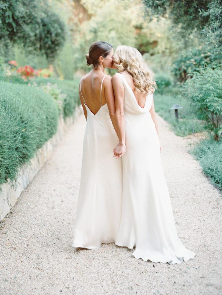 In questa foto due spose riprese di spalle si baciano mentre camminano sul viale di un giardino con i siepi e alberi dopo la loro unione civile. Entrambe indossano un abito da sposa scivolato in chiffon di sera. L'abito della sposa a sinistra, mora con i capelli raccolti in un chiffon, ha delle bretelle sottilissime con la schiena scoperta. La sposa a destra, con i capelli biondi sciolti e mossi, indossa un abito con una scollatura morbida a sacchetto sulla schiena  