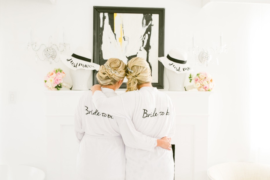 In questa foto due spose bionde di spalle con i capelli raccolti prima delle loro nozze. Indossano una vestaglia bianca con una scritta nera sulla schiena che riporta la frase "Bride to be". Sulla mensola sono poggiati i loro bouquet di fiori rosa e bianchi e due cappelli bianchi a tesa larga con una fascia nera e una scritta nera con la frase "Just married"