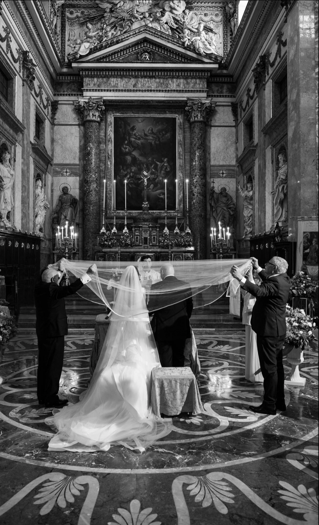 In questa foto in bianco e nero due sposi di spalle durante il rito della velazione. Sopra le loro teste è disteso un velo tenuto agli angoli dai quattro genitori