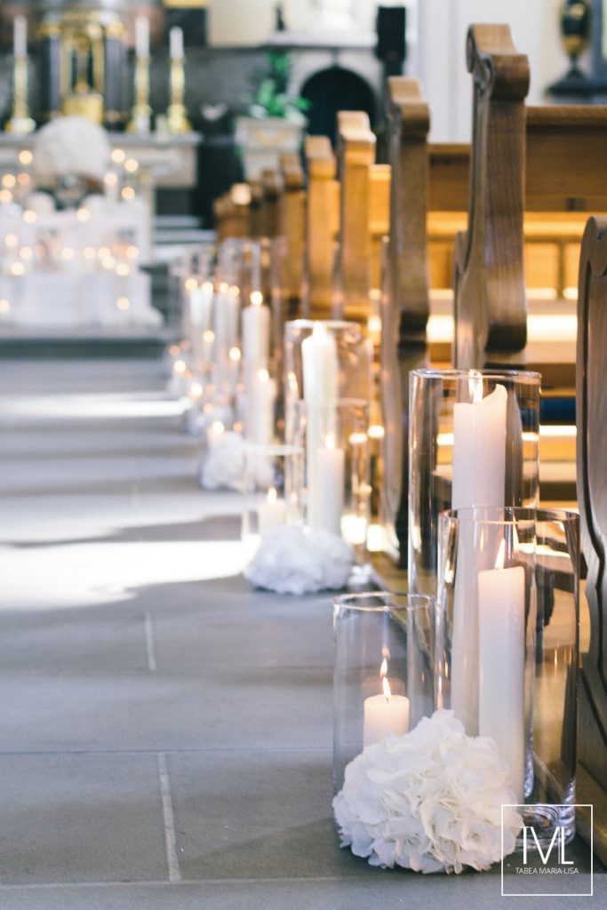 In questa foto una chiesa decorata lungo la navata con candele poggiate sul pavimento e accese in cilindri trasparenti e sfere di fiori bianchi