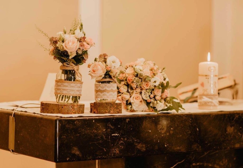 In questa foto una mensa della chiesa decorata in stile shabby chic con vasi di vetro decorati con nastri di juta e pizzo poggiati su cilindri di legno. I fiori scelti sono rose bianche e rosa