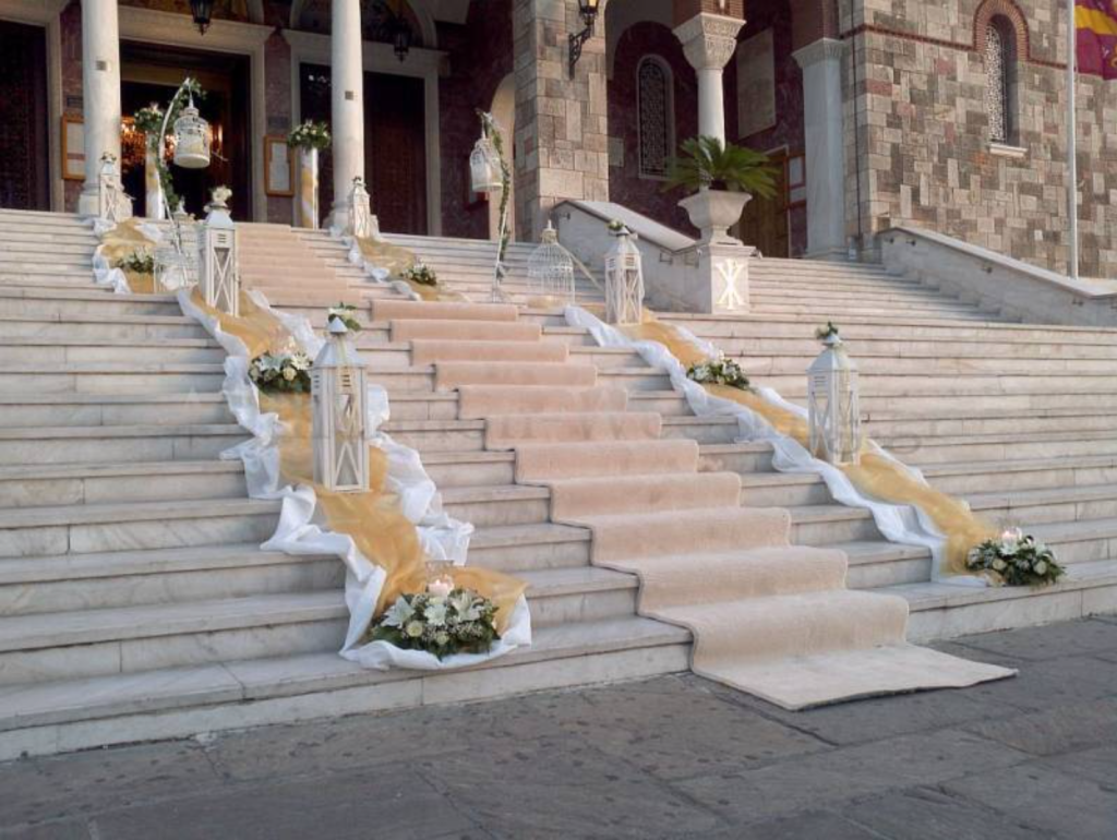 In questa foto i gradini di una chiesa decorati con un tappeto colore avorio, lanterne, tulle colore avorio, drappi di stoffa bianca e composizioni di fiori abbinati