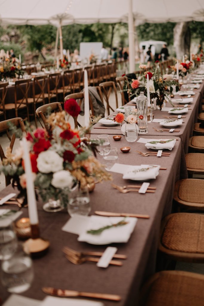 In questa foto gli allestimenti matrimonio per un ricevimento autunnale realizzato con tavoli imperiali con tovagliato marrone, piccoli centrotavola di fiori bianchi, rossi e gialli, posate oro rosa e rametti di erbe aromatiche usati come segnaposto 