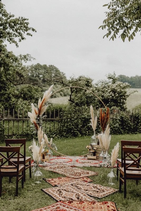 In questa foto l'allestimento di una cerimonia civile in stile Boho Chic organizzata su un prato. Lungo il corridoio centrale sono disposti tappeti fino all'altare realizzato con un cerchio decorato con pampas. Lungo il corridoio sono disposti vasi trasparenti con pampas beige disposti accanto a sedie di legno scuro