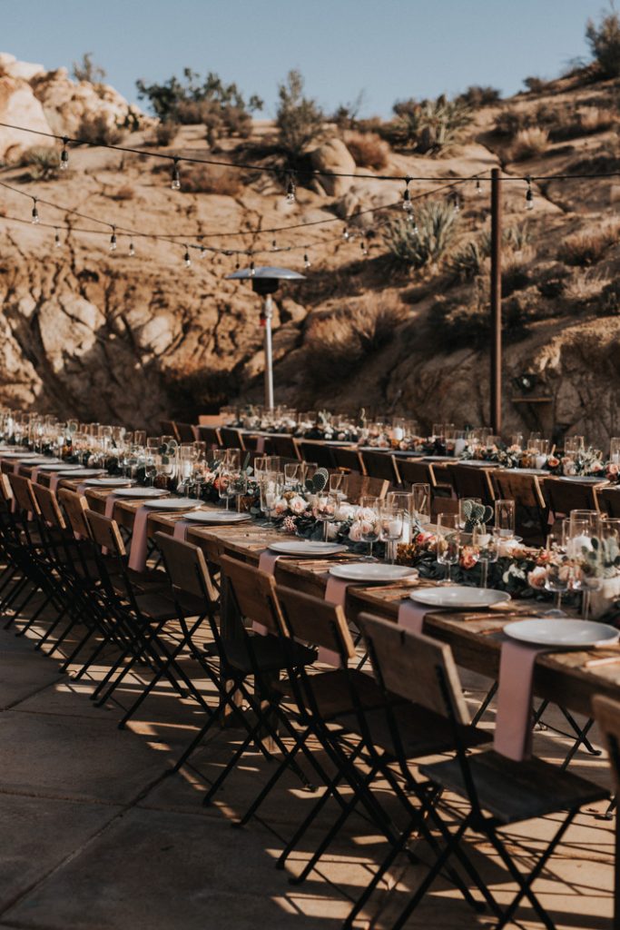 In questa foto gli allestimenti matrimonio di un ricevimento nel deserto in stile country chic nei toni del rosa cipria e dell'arancione. Nella foto sono ripresi in obliquo due tavoli imperiali di legno con sedie rustiche. Al centro dei tavoli è disposto un lungo runner di fiori di campo con portacandele trasparenti